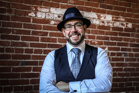 Drew Dudley wears a fedora, a dress shirt, vest and tie. He is standing in front of a brick wall and smiling.